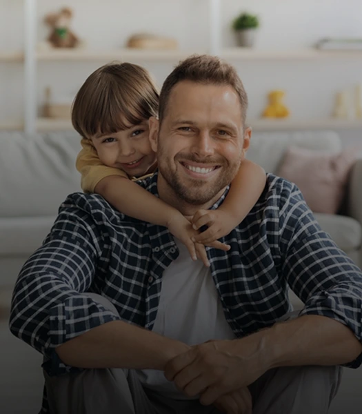 A smiling father sitting on the floor with his young child hugging him from behind, representing paternity or father-child relationship.