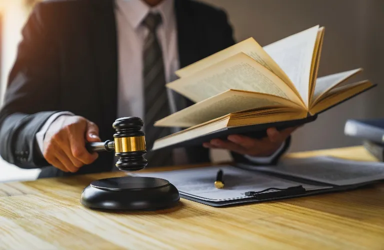 Person in formal suit holding a gavel and open book at a wooden desk with documents.