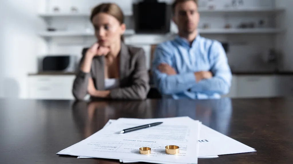 Two gold wedding rings on divorce papers with a pen, blurred couple in background.