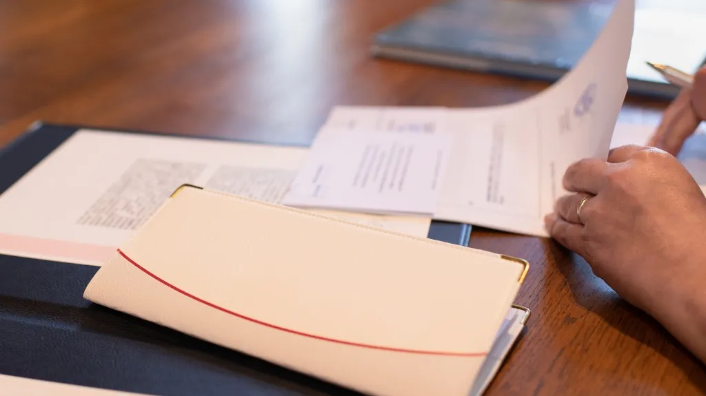 Person's hands reviewing documents on a wooden table with a white folder and pen.