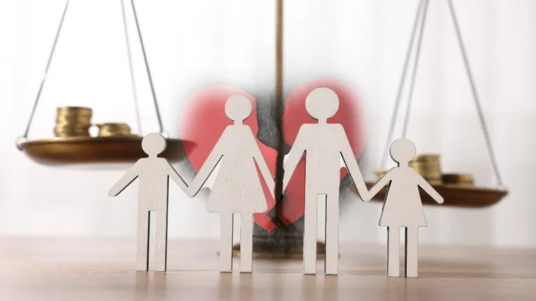 Wooden cutout figures of a family holding hands in front of a broken red heart and balancing scales with coins.