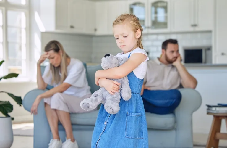 Young girl in blue dress holding teddy bear, parents seated and distressed in background.
