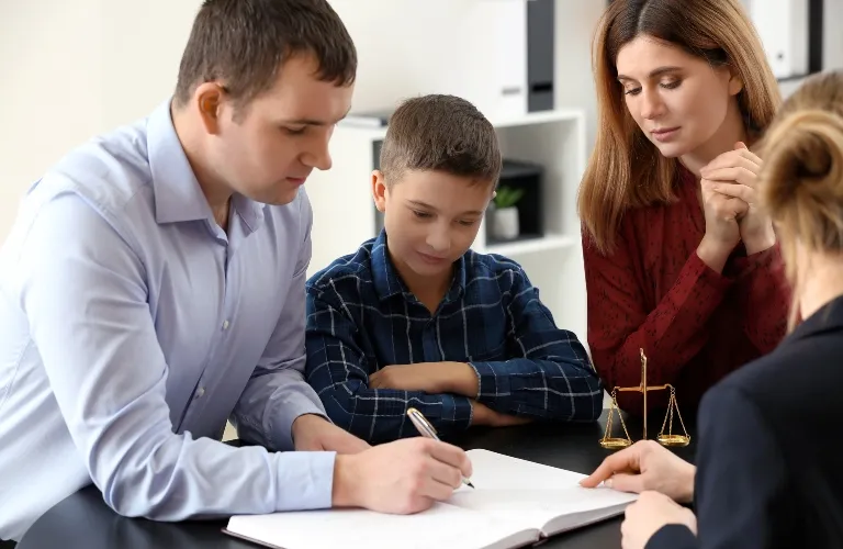 Four people, including a man signing a document, a boy, and two women, seated at a table with legal scales.