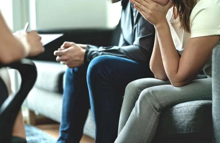 Two individuals seated on a gray couch, one covering their face with hands, the other with clasped hands, in a counseling session.