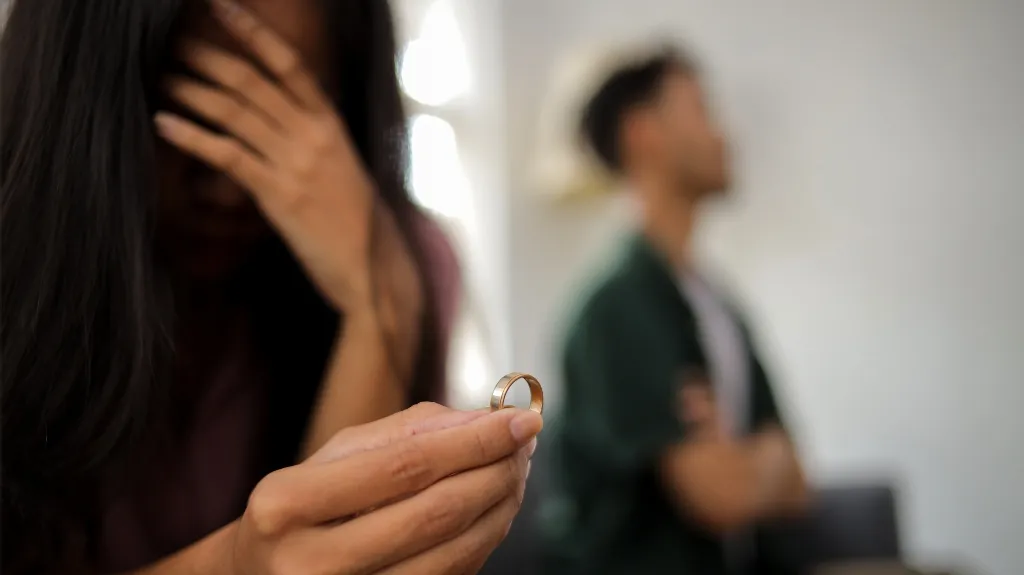 Woman holding a wedding ring with distressed expression, man blurred in background.