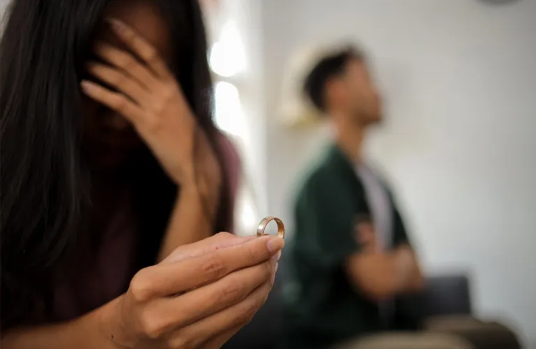Woman holding a wedding ring, covering her face, man sitting in background with crossed arms.