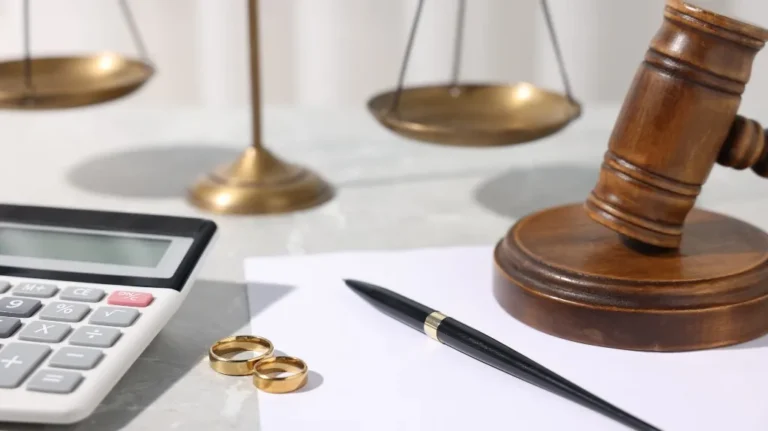 Wooden judge's gavel, gold wedding rings, calculator, pen, and brass scales on white surface.