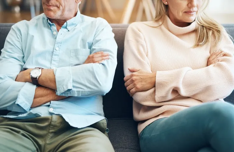 Middle-aged man and woman sitting on a couch, arms crossed, appearing upset.