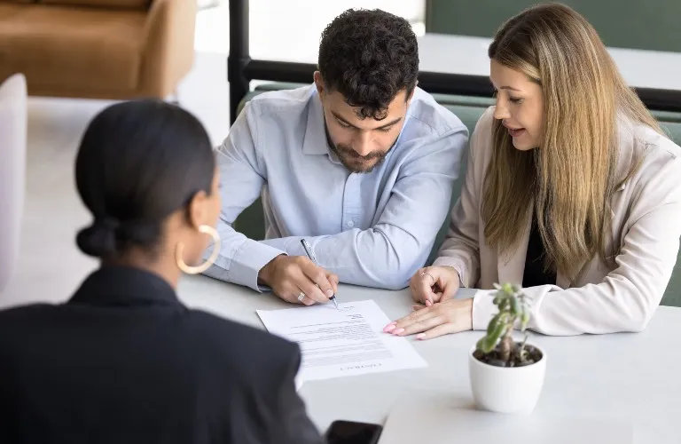 Three professionals at a table, a man signing a contract document while two women observe.
