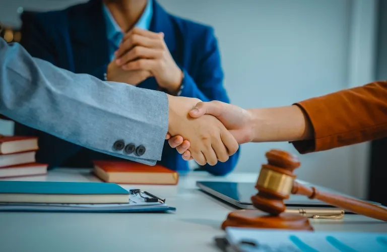 Two individuals shaking hands across a desk with legal books, a gavel, and documents, while a third person in a blue suit observes.