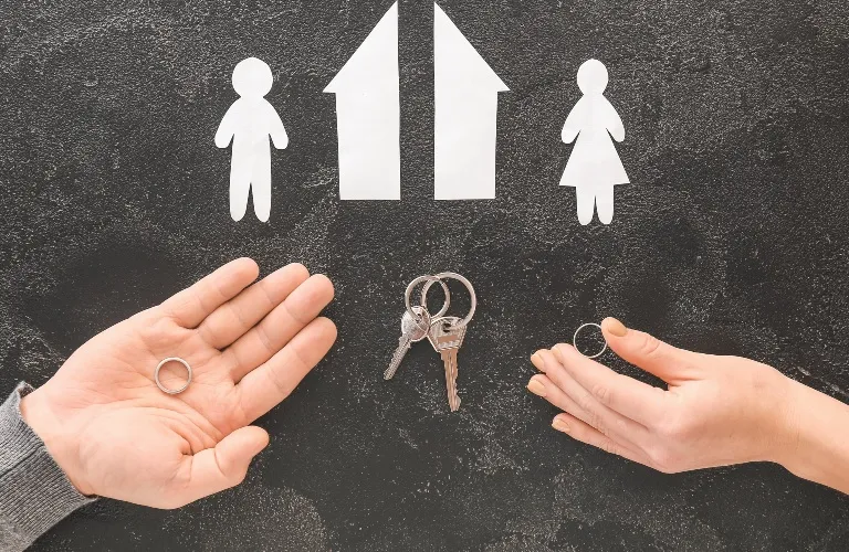 Two hands holding wedding rings beneath white paper cutouts of a man, woman, house, and keys on a black surface.