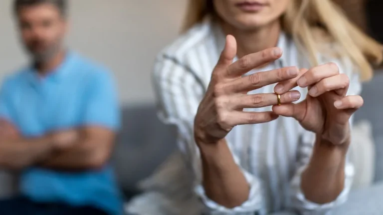 Woman removing gold wedding ring from finger with blurred man in blue shirt sitting in background.