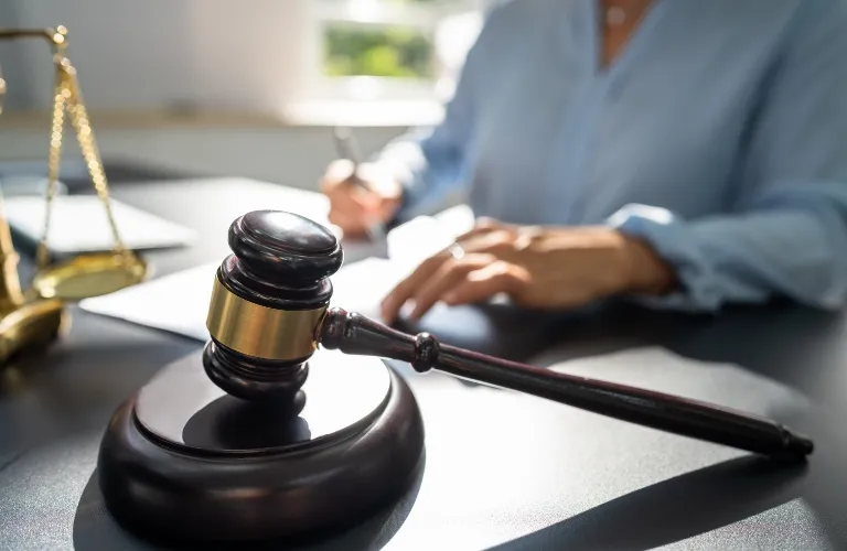 Close-up of a dark wooden gavel on a desk with a blurred person writing in the background.