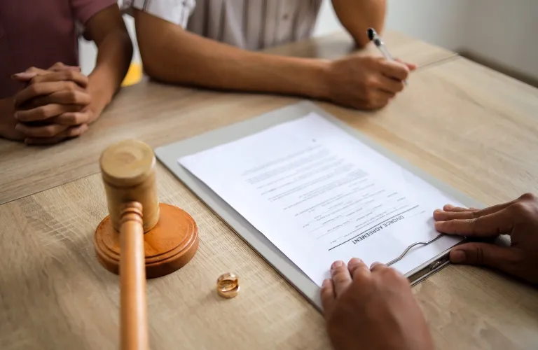 Close-up of a legal document, wooden gavel, wedding rings, and people signing papers at a table.