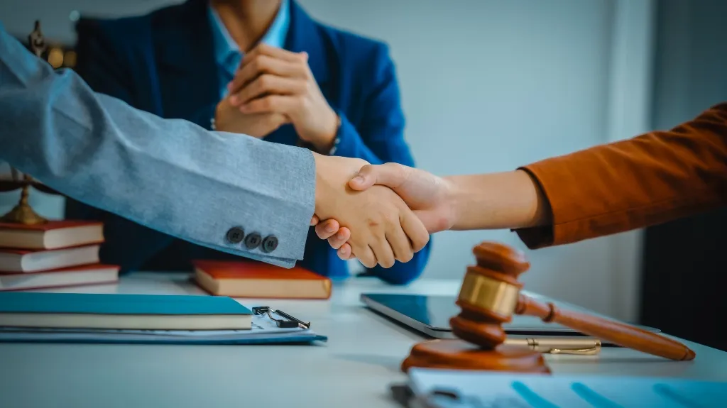 Two individuals in business attire shaking hands over a desk with legal books, documents, and a wooden gavel.