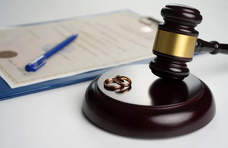 Wooden judge's gavel with two gold wedding rings on base, blurred document and blue pen in background.