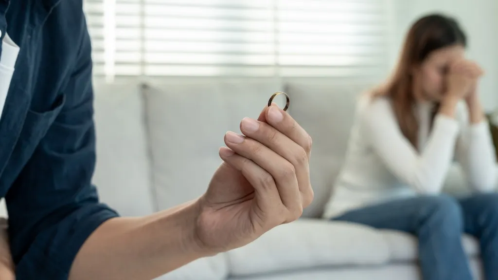 Man holding a wedding ring with a distressed woman sitting on a couch in the background.