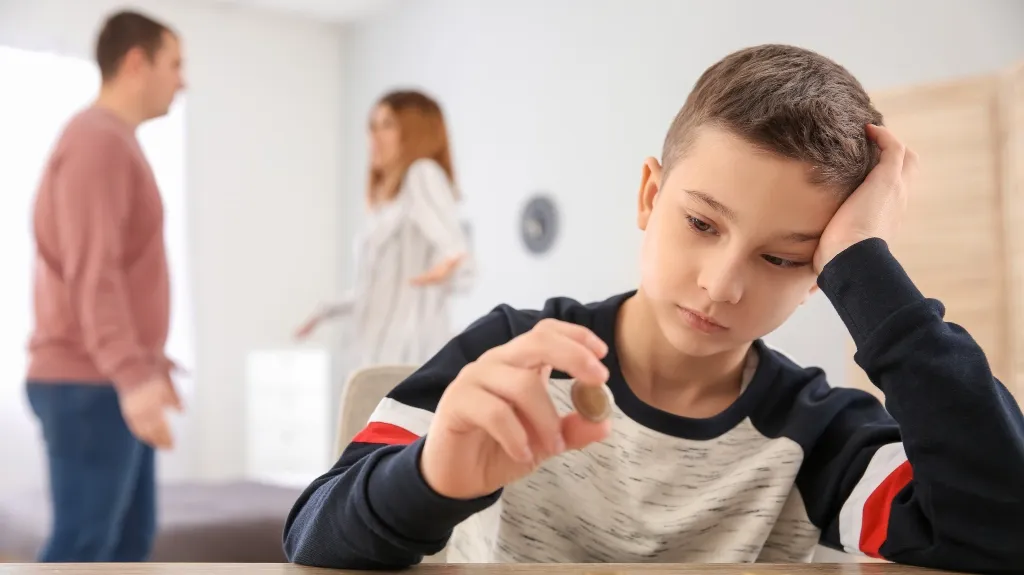 Young boy looking thoughtfully at a coin while parents argue in the background.