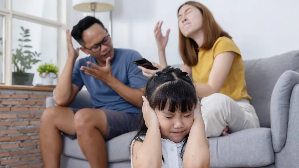 Young girl covering ears with eyes closed, parents arguing on gray sofa behind her.
