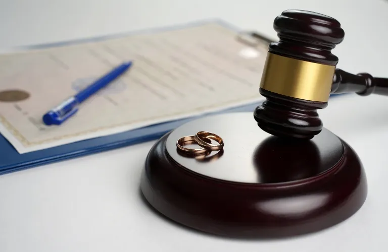 Wooden judge's gavel with two gold wedding rings and a legal document with blue pen on white surface.