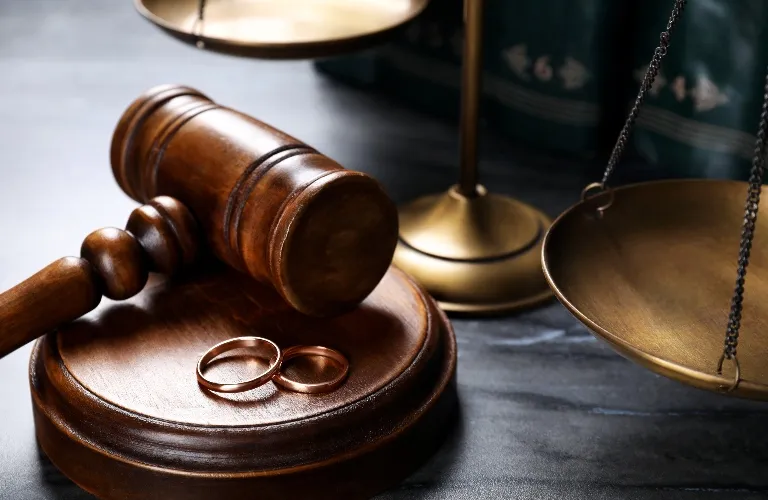 Wooden judge's gavel with two gold wedding rings and brass scales of justice on dark surface.