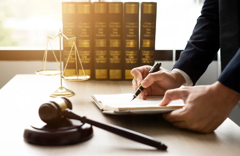 Person in dark suit writing on a document at a desk with legal books, scales of justice, and gavel.