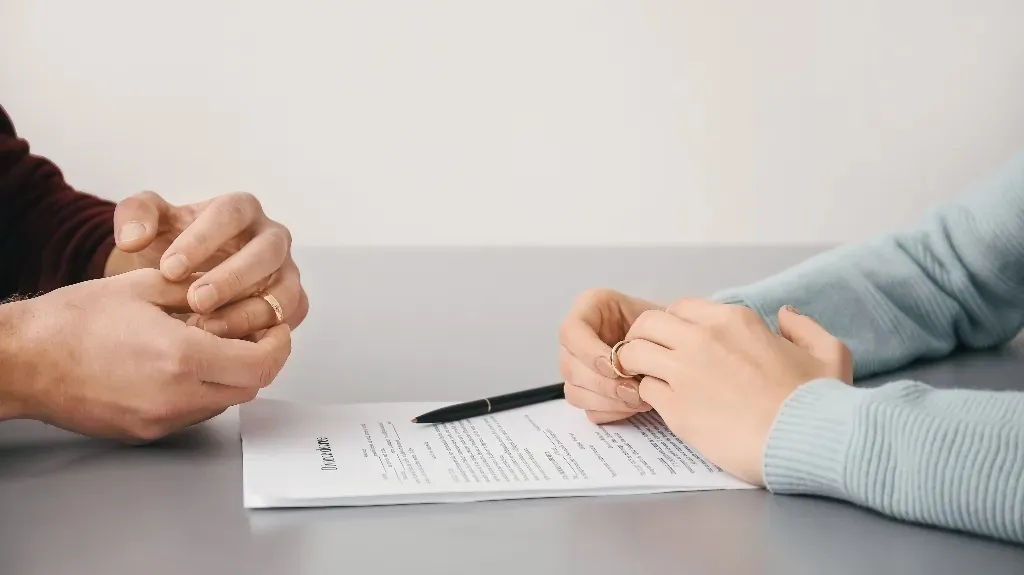 Two individuals sitting at a table, holding wedding rings over a divorce agreement document.