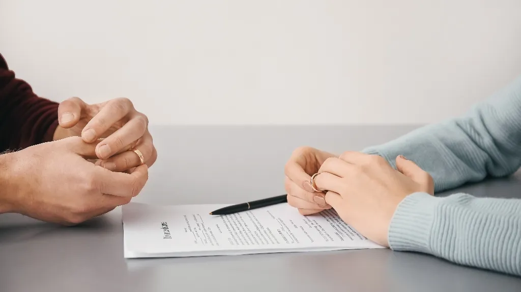 Two individuals holding wedding rings over a divorce agreement document on a table.