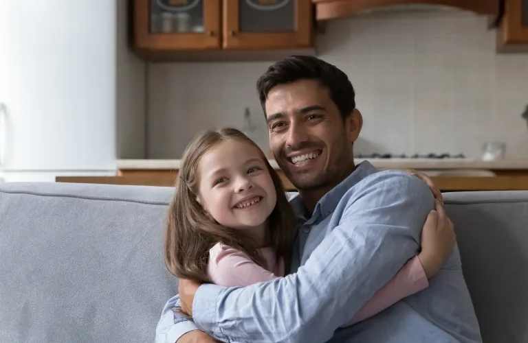 Man and young girl smiling and embracing on a gray couch in a modern kitchen.