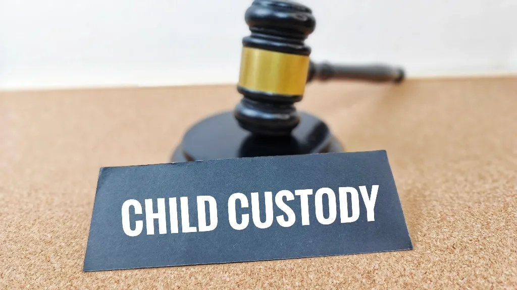 Black and gold judge's gavel behind a black sign with white text "CHILD CUSTODY" on corkboard.