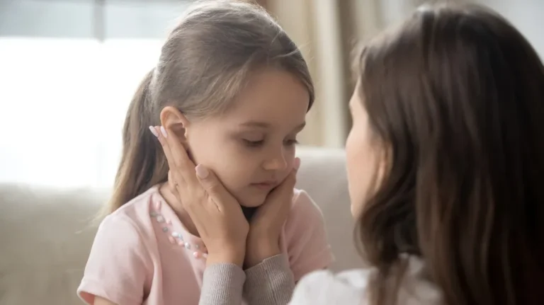 Woman gently holding a young girl's face, both with neutral expressions indoors.